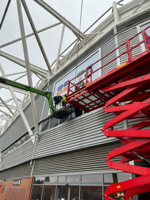 Two team members stand atop a red crane outside Southampton Football Club Stadium, Southampton