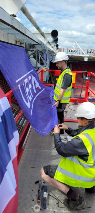 Two members of the team at Elmtree Signs place flags to the roof of a stadium